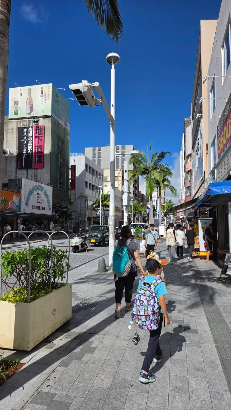 家人背著背包走在國際通街道上，兩旁是商店與棕櫚樹